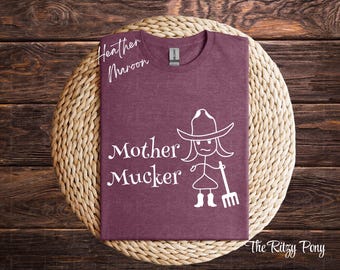May include: Heather maroon t-shirt with the words "Mother Mucker" and a stick figure wearing a cowboy hat and holding a pitchfork. The shirt is displayed on a woven circular mat.