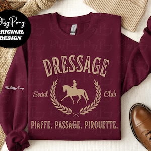 Pu&ograve; includere: Felpa bordeaux con la scritta "DRESSAGE Social Club" in stile vintage. Il design include una silhouette di cavallo e cavaliere all'interno di una corona d'alloro. Le parole "PIAFFE. PASSAGE. PIROUETTE." sono stampate sotto.