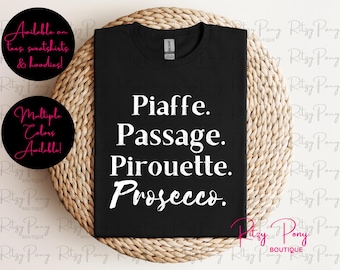 May include: Black t-shirt with white text listing ballet terms: Piaffe, Passage, Pirouette, and Prosecco. The shirt is displayed on a woven surface. Additional text on the image indicates the shirt is available in multiple colors.