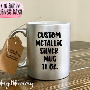 May include: A silver metallic mug with the text "CUSTOM METALLIC SILVER MUG 11 OZ." printed in black. The mug has a matching silver handle and a tag attached with twine. The mug is on a wooden surface.