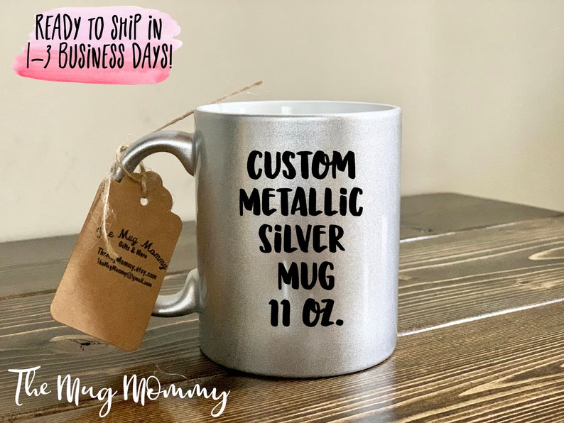 May include: A silver metallic mug with the text "CUSTOM METALLIC SILVER MUG 11 OZ." printed in black. The mug has a matching silver handle and a tag attached with twine. The mug is on a wooden surface.