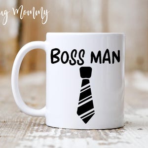 May include: A white ceramic mug featuring the black text BOSS MAN printed in a bold font above a black and white graphic illustration of a necktie. The mug has a standard handle on the left side and a smooth, glossy finish on the exterior surface.