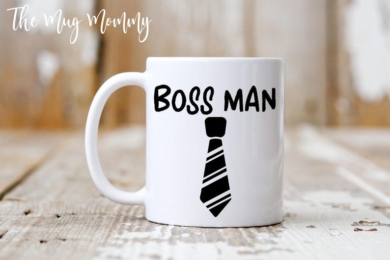 May include: A white ceramic mug featuring the black text BOSS MAN printed in a bold font above a black and white graphic illustration of a necktie. The mug has a standard handle on the left side and a smooth, glossy finish on the exterior surface.