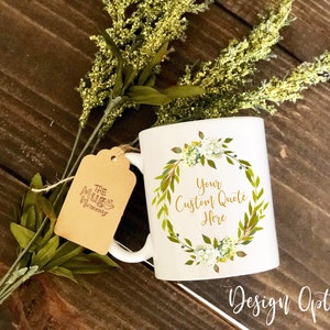 K&ouml;nnte beinhalten: Ein wei&szlig;er Keramikbecher mit einem aufgedruckten gr&uuml;n-gelben Blumenkranz-Design und dem Text Your Custom Quote Here in der Mitte. Ein braunes Papieretikett mit dem Text The Mug Mommy ist mit einer d&uuml;nnen Schnur am Henkel befestigt.