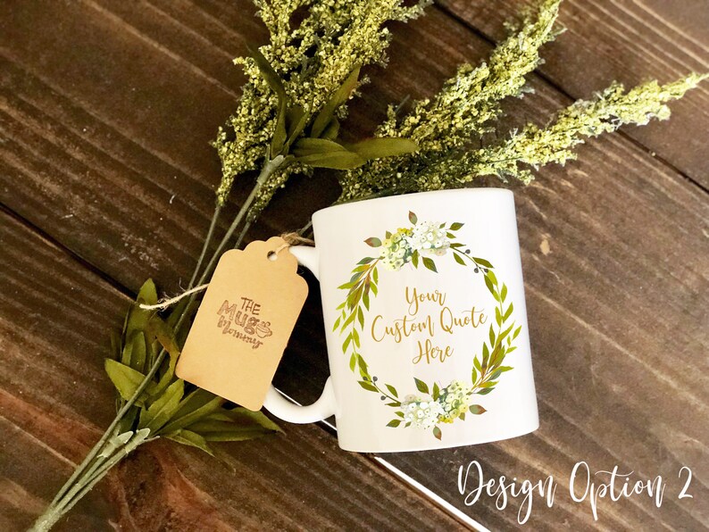 K&ouml;nnte beinhalten: Ein wei&szlig;er Keramikbecher mit einem aufgedruckten gr&uuml;n-gelben Blumenkranz-Design und dem Text Your Custom Quote Here in der Mitte. Ein braunes Papieretikett mit dem Text The Mug Mommy ist mit einer d&uuml;nnen Schnur am Henkel befestigt.