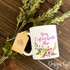 K&ouml;nnte beinhalten: Ein wei&szlig;er Keramikbecher mit dem violetten Text "Your Custom Quote Here" &uuml;ber einem bunten Blumenarrangement. Ein braunes Papieretikett mit dem Logo "The Mug Mommy" ist mit einer Schnur am Henkel befestigt, und gr&uuml;nes Blattwerk liegt hinter dem Becher.