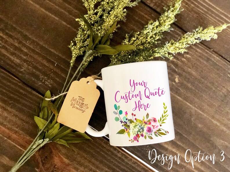K&ouml;nnte beinhalten: Ein wei&szlig;er Keramikbecher mit dem violetten Text "Your Custom Quote Here" &uuml;ber einem bunten Blumenarrangement. Ein braunes Papieretikett mit dem Logo "The Mug Mommy" ist mit einer Schnur am Henkel befestigt, und gr&uuml;nes Blattwerk liegt hinter dem Becher.
