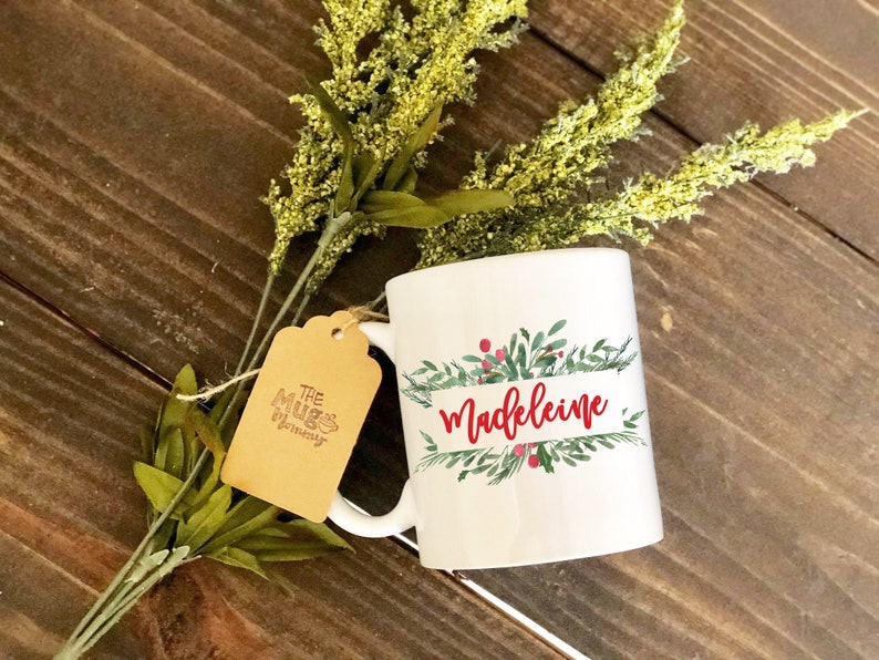 May include: White ceramic mug with the name "Madeleine" in red script, surrounded by a green and red floral design. A brown tag with "The Mugs Mommy" is attached. Green and yellow floral stems are in the background.