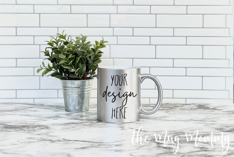 May include: A silver mug with a white interior, featuring the text "YOUR design HERE" in black. A small potted plant sits to the left of the mug. The mug is on a marble-patterned surface against a white brick wall.