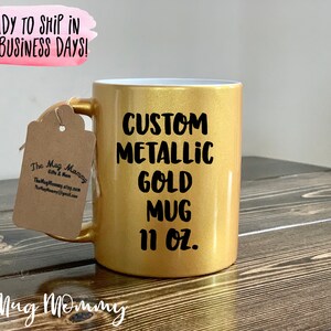 May include: A custom metallic gold mug with the text "CUSTOM METALLIC GOLD MUG 11 OZ." printed in black. The mug has a gold handle and a tag attached. The mug is on a wooden surface.