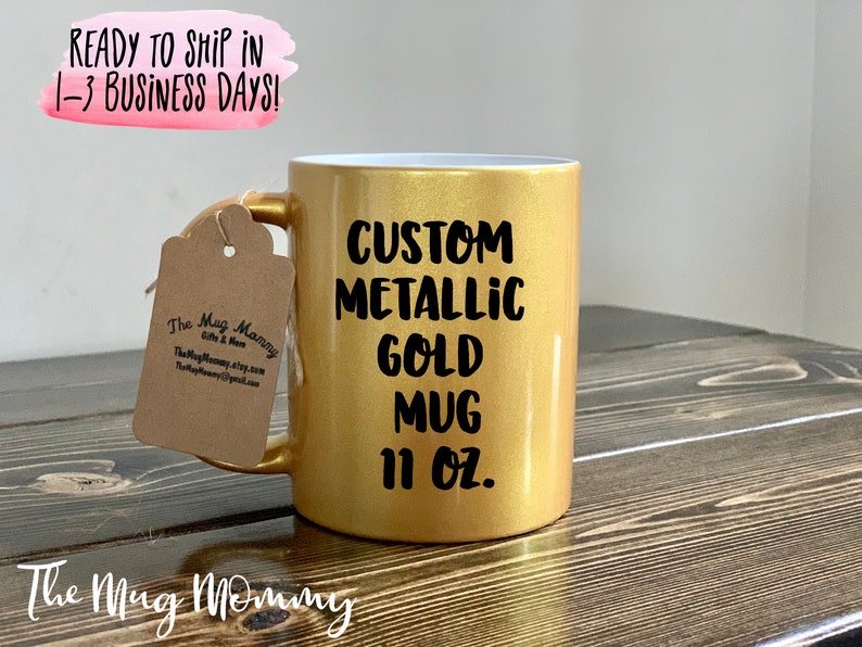 May include: A custom metallic gold mug with the text "CUSTOM METALLIC GOLD MUG 11 OZ." printed in black. The mug has a gold handle and a tag attached. The mug is on a wooden surface.