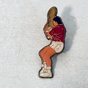 May include: Vintage tennis player pin with a gold-tone border. The pin features a figure in a red shirt, white shorts, and white shoes, holding a tennis racket. The figure is in a playing position.