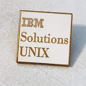 Può includere: Una spilla quadrata con sfondo bianco e bordi dorati. La spilla presenta il testo "IBM Solutions UNIX" in lettere dorate. Il logo IBM è nella parte superiore della spilla.