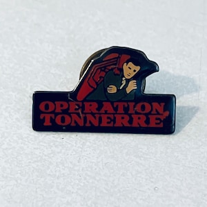 May include: A black and red enamel pin with a cartoon illustration of a man with a red bird on his shoulder. The text "OPERATION TONNERRE" is printed in red on a black background.