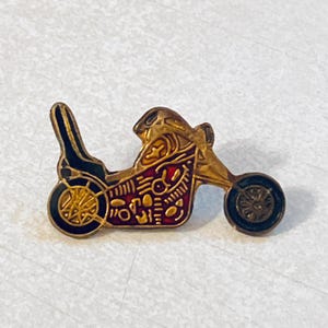 Op de afbeelding: Een vintage motorfietsspeld met gouden, zwarte en rode emaille details. De speld heeft een gestileerd ontwerp van een motorfiets met ingewikkelde patronen en een klassieke esthetiek. De wielen en de carrosserie zijn gedetailleerd.