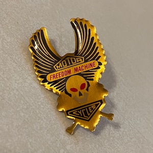 Op de afbeelding: Een goudkleurige metalen pin met een schedel- en vleugelontwerp. De pin bevat de woorden "MOTOR FREEDOM MACHINE" in rood en zwart. De pin heeft ook het woord "CYCLES" in een banner aan de onderkant.