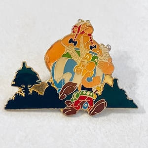 Puede incluir: Un colorido pin de esmalte con un personaje de dibujos animados con pelo naranja, una túnica a rayas azules y blancas y pantalones rojos. El pin tiene un borde dorado y una silueta verde oscuro de árboles en el fondo.