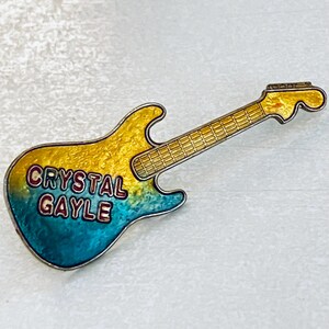 May include: A guitar-shaped pin with a gradient of yellow to blue enamel. The pin features the text "CRYSTAL GAYLE" in red lettering. The guitar's neck and headstock are gold-colored.