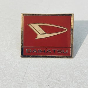 Op de afbeelding: Een gouden speld met een rode achtergrond en een gestileerde witte letter D. De tekst "DAIHATSU" is in gouden letters onder het logo gedrukt.