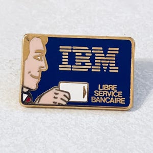 Op de afbeelding: Een vintage IBM-speld met een gouden rand. De speld heeft een blauwe achtergrond met het IBM-logo in goud. Een cartoonfiguur houdt een witte kaart met een pijl vast. De tekst "LIBRE SERVICE BANCAIRE" is ook aanwezig.