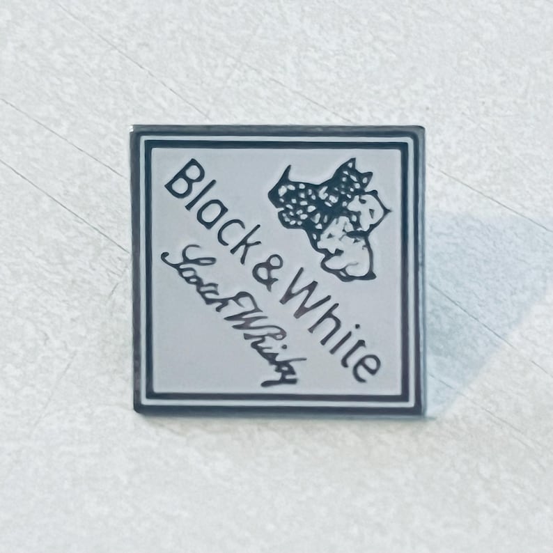 May include: Square, silver-colored pin with a black border. The pin features the words "Black & White" in a bold font, with "Scotch Whisky" written below in a cursive script. A black and white graphic of a dog is in the upper right corner.