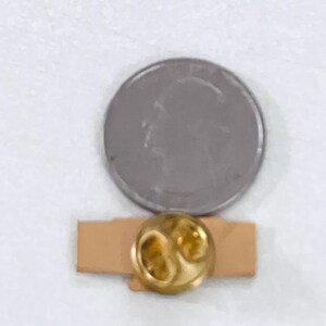 May include: A gold-colored lapel pin with a unique design, set on a small, rectangular, tan-colored backing. A silver coin is next to the pin for size comparison. The pin has a shiny, reflective surface.