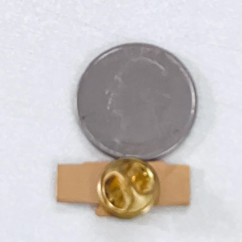 May include: A gold-colored lapel pin with a unique design, set on a small, rectangular, tan-colored backing. A silver coin is next to the pin for size comparison. The pin has a shiny, reflective surface.