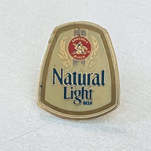以下が含まれることがあります： 盾形の金属製ピンバッジで、ゴールド調の背景にダークブルーの「Natural Light BEER」の文字が記されています。文字の上部には、アンハイザー・ブッシュのイーグルとAのシンボルが入った赤い円形ロゴがあり、その両脇には金色の麦の穂が描かれています。