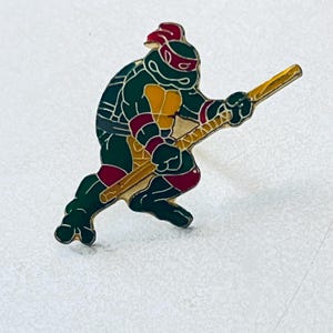 May include: A metal enamel pin featuring a green turtle character wearing a red mask and elbow pads, a yellow chest plate, and a brown shell. The character is in a dynamic pose, holding a long, light brown wooden staff extended diagonally across the body.