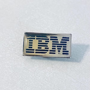 Puede incluir: Un pin rectangular con las letras "IBM" en azul sobre un fondo dorado. El pin tiene un borde plateado y es probablemente un artículo vintage o de colección, posiblemente un logotipo corporativo o un artículo promocional.