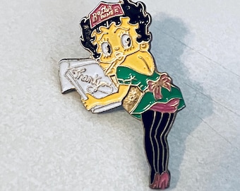 Op de afbeelding: Kleurrijke emaille pin met een stripfiguur in een groene jurk en zwarte kousen, met een bord met de tekst "Bedankt". De pin heeft een "Betty Diner" logo op het hoofd van het personage. Een verzamelobject.