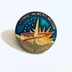 May include: A round, enameled pin with a blue and gold design. The pin features the text "LOUNGE HILMERS NELSON" and "HAUCK COVEY". The design includes a stylized starburst and a red ribbon.