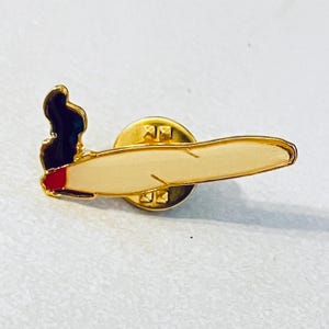 May include: A gold-coloured pin badge in the shape of a lit cigarette. The badge has a tan body with a red tip and black smoke detail. The pin is attached to a gold-coloured backing.