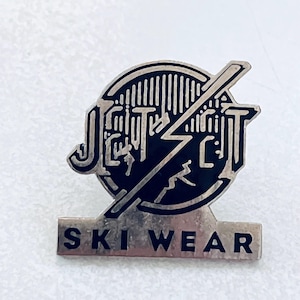 May include: A silver-toned pin with the words "Jetric" and "Ski Wear". The pin features a black circle with a lightning bolt and a skier silhouette. The text is in a stylized font.