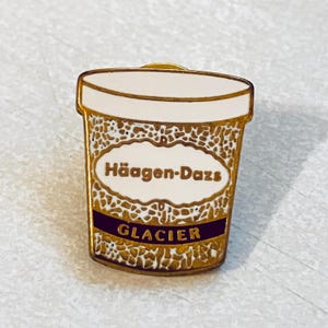 May include: A gold-colored enamel pin in the shape of a Häagen-Dazs ice cream container. The pin features the brand name and the word "GLACIER" in a dark purple banner. The container has a white top and a textured gold pattern.