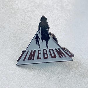 Puede incluir: Un pin de esmalte plateado con un diseño triangular. El pin presenta una silueta negra de dos figuras corriendo, sobre un fondo blanco. La palabra "TIMEBOMB" está escrita en letras rojas en la base del triángulo.