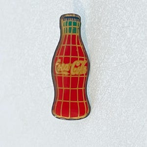 May include: A vintage Coca-Cola bottle-shaped pin. The pin features a red background with a gold grid pattern and the Coca-Cola logo in gold. The bottle cap is green and the edges are gold.