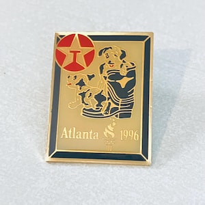 May include: A gold-coloured pin featuring a red star with the letter "T" inside. Two black and white cartoon dogs are in the centre of the pin, with the text "Atlanta 1996" below them.