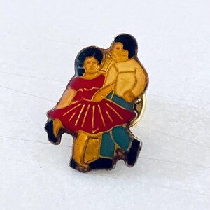 Op de afbeelding: Een vintage emaille speld met een dansend stel. De speld toont een man en een vrouw in een danshouding, met de vrouw in een rode jurk en de man in een geel shirt en een blauwe broek. De speld heeft een goudkleurige rand.