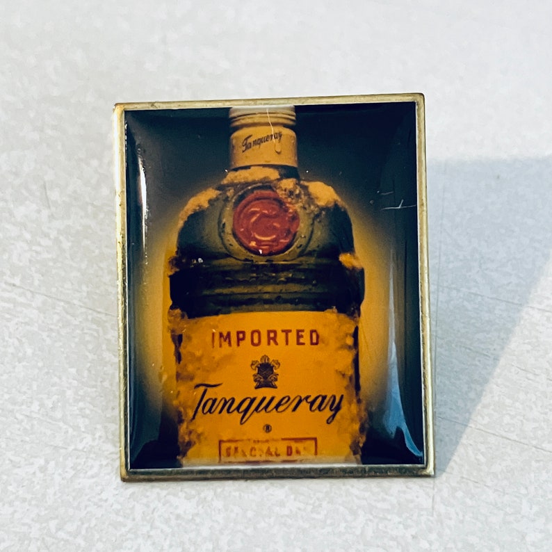 May include: A gold-colored metal pin with a frosted glass bottle of Tanqueray gin. The bottle is partially covered in frost and has a red wax seal. The text "IMPORTED Tanqueray" is printed on the bottle.