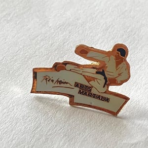 May include: A vintage pin featuring a martial artist in a karate stance. The pin has a gold-colored border and a white background with the words "ARTS MARTIAUX" and a signature. The martial artist is wearing a white uniform with orange and black accents.