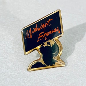 May include: A gold-toned pin featuring a black rectangle with the words "Midnight Express" in orange script. Below the rectangle is a black silhouette of a person looking upward. The pin has a gold border.