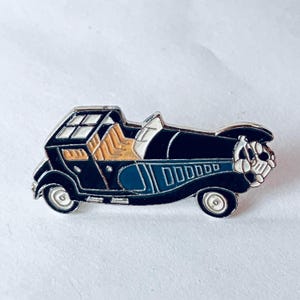 May include: A vintage-style car pin with a black body, blue accents, and silver wheels. The pin features a detailed design with a white top and tan interior. The car has a classic, antique appearance.