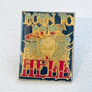 以下が含まれることがあります： ダークブルーの背景に、上部にゴールドの文字で「BORN TO RAISE」、下部に「HELL」と記された長方形のエナメルピン。中央には、小さな赤い点がある様式化された顔のゴールドトーンのグラフィックが描かれています。