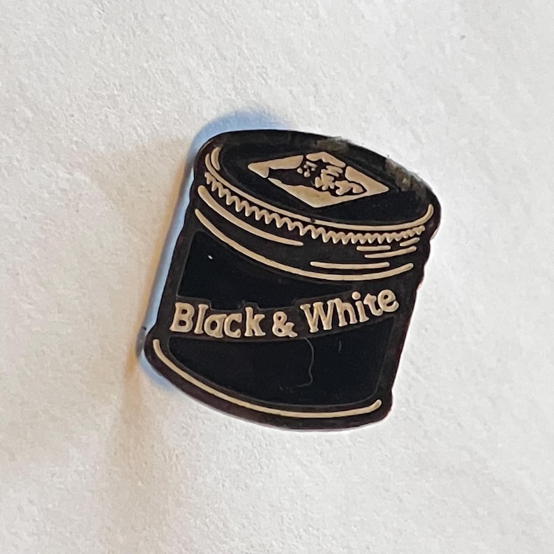 May include: A black and white pin shaped like a can, with the words "Black & White" in a banner across the front. The top of the can is black with a white zig-zag design. The pin is on a white background.