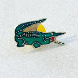 Op de afbeelding: Een metalen emaille speld in de vorm van een krokodil in profiel, met een donkergroen lichaam met een gestructureerd schubbenpatroon, een lichtbruine buik en een klein rood accent in de geopende bek tegen een gele achtergrond.