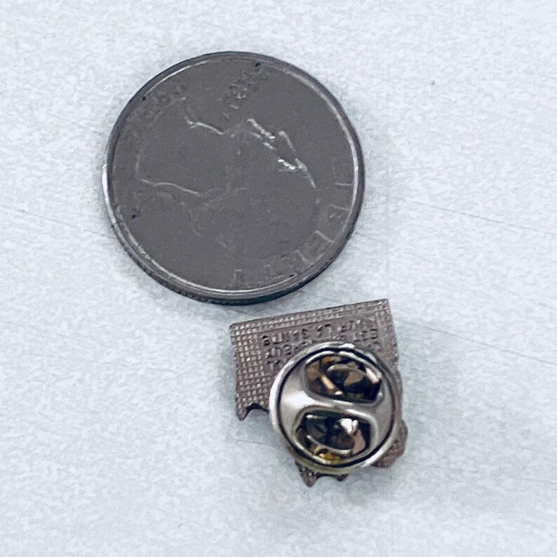 Puede incluir: Una moneda de veinticinco centavos de color plateado junto a un peque&ntilde;o pin rectangular. El pin tiene un borde plateado y un cierre de alfiler. El texto en el pin es dif&iacute;cil de leer. El pin es probablemente un objeto de colecci&oacute;n o un recuerdo.