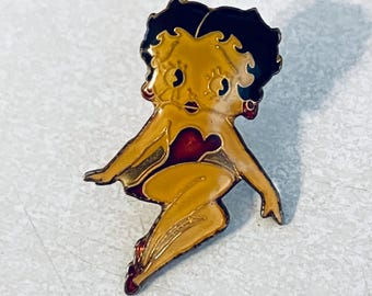 Op de afbeelding: Een vintage emaille speld met een stripfiguur in een zittende pose. De speld heeft een gele body, zwart haar en een rood hartvormig detail. De speld is goudkleurig omlijnd en heeft rode accenten.