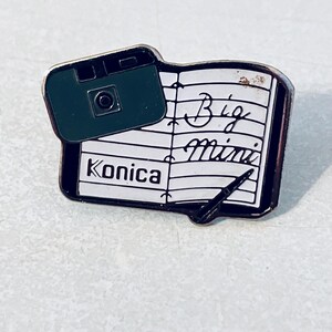 May include: Black and white enamel pin featuring a vintage Konica camera and a notebook with the words "Big Mini" written on it.