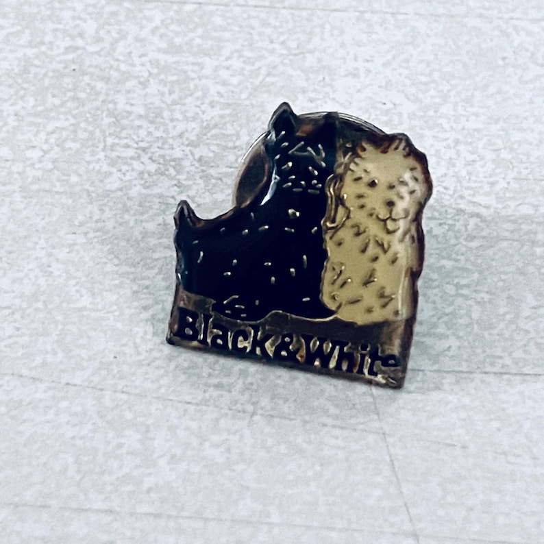 Puede incluir: Un pin rectangular con dos retratos estilizados de perros, uno negro y otro beige, con las palabras "Black & White" en la parte inferior. El pin tiene un borde met&aacute;lico y un cierre de alfiler.